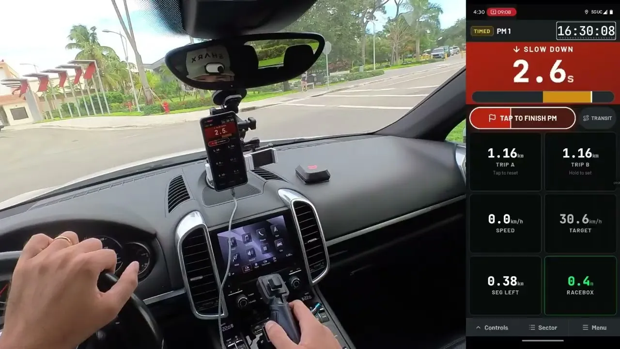 Inside a car: a phone on the dashboard showing Crono107’s average-speed trial, the navigator’s hand on a handheld remote and a RaceBox Mini on the dash, with the app screen enlarged beside it