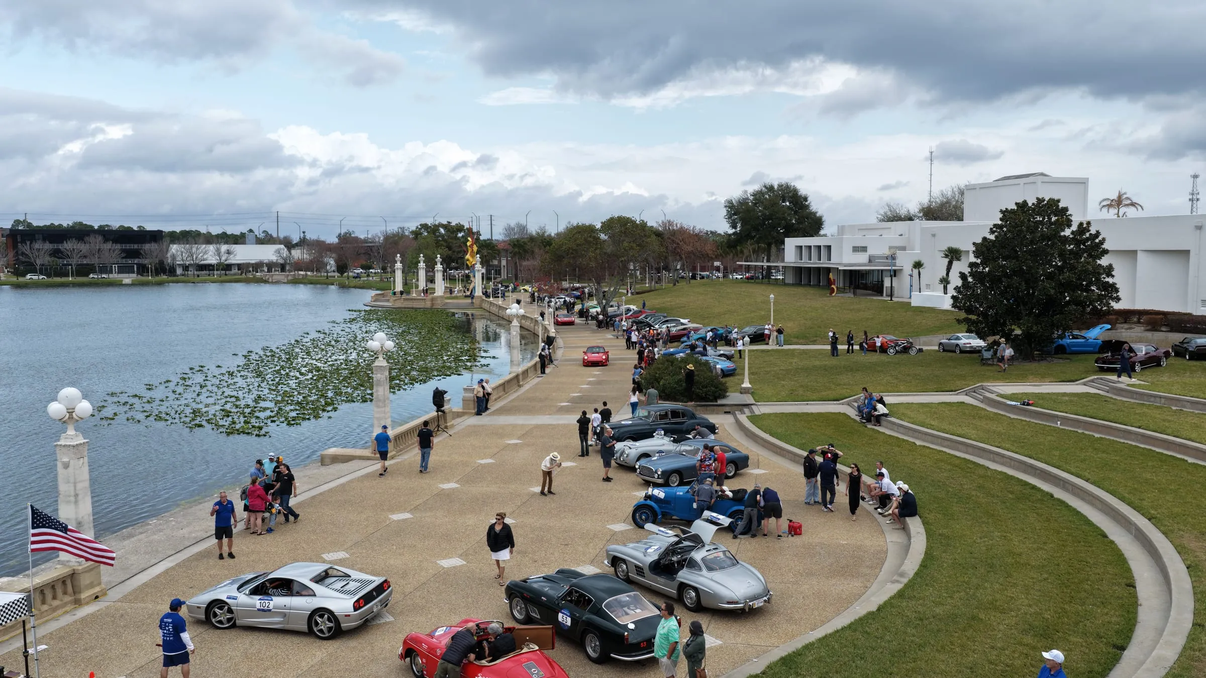 Classic rally cars lined up along a lakeside promenade between stages, with crews and spectators around them