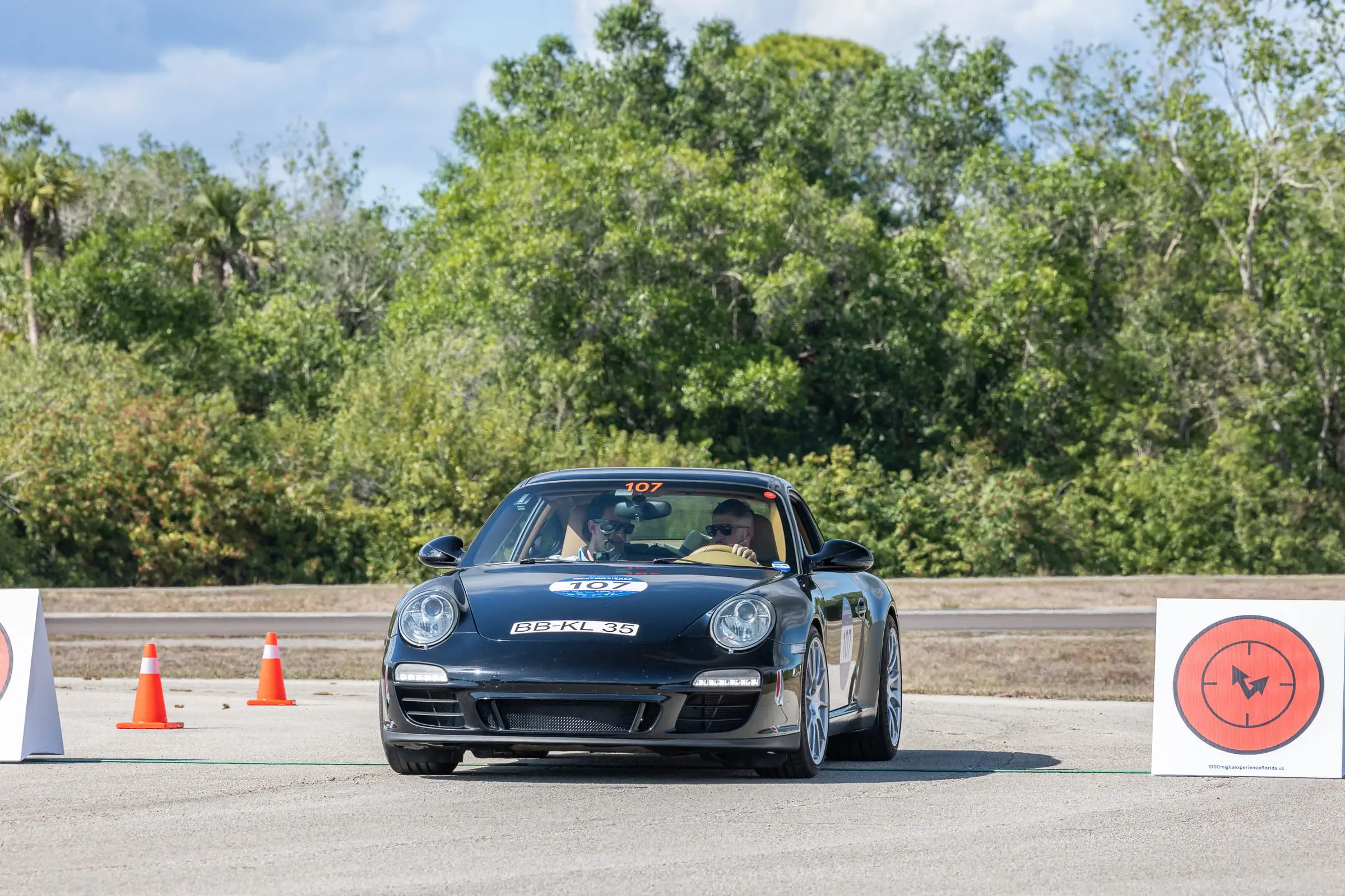 Car 107, a black Porsche 911, crossing the line at a timed trial sign between cones, with the crew inside