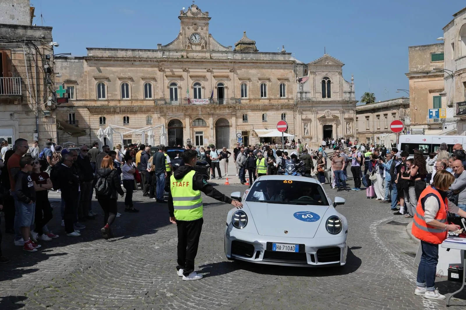 Car 60 arriving in a crowded square in front of a baroque palace, with a marshal waving it in