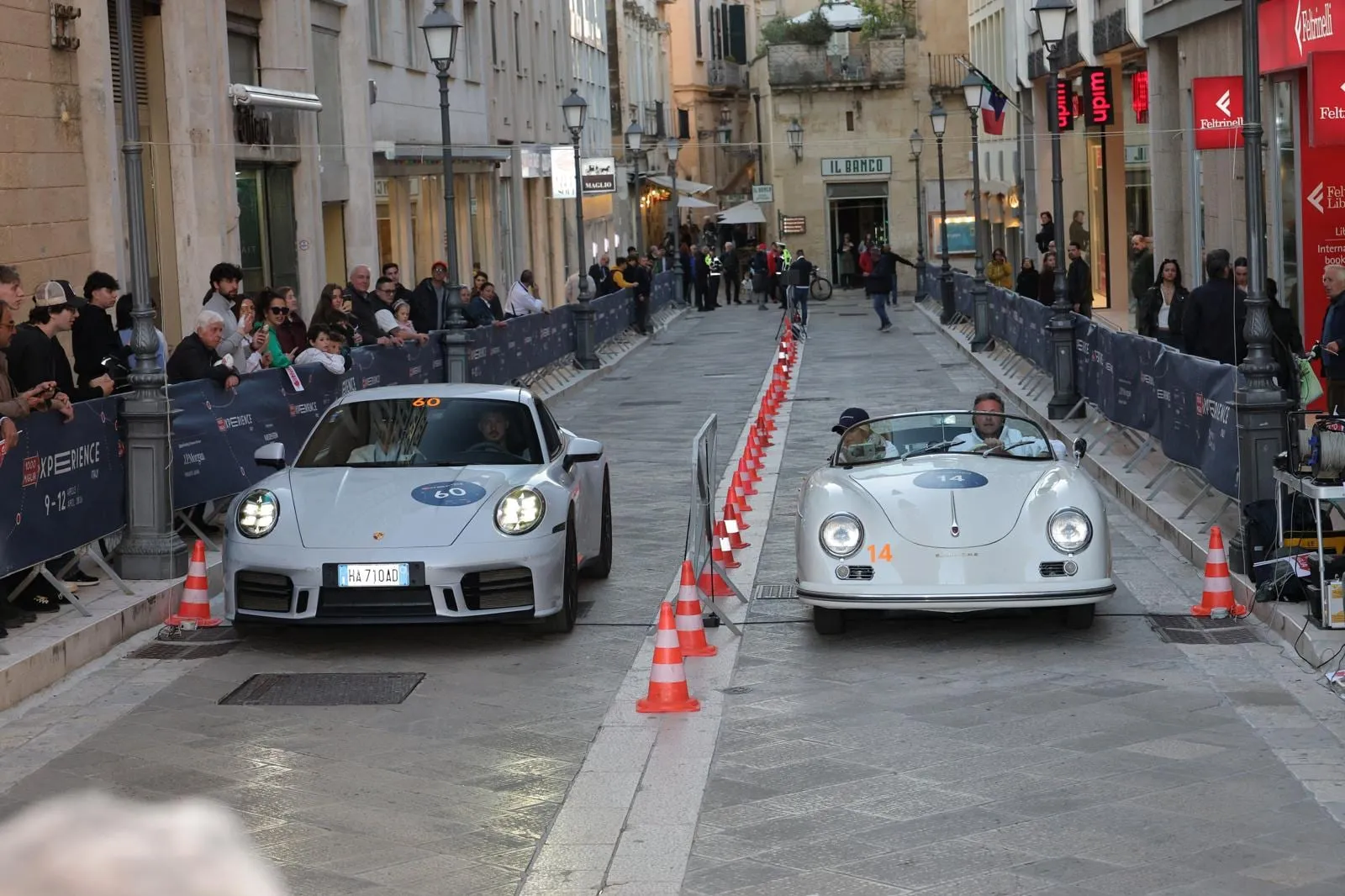 Car 60, a silver 2025 Porsche 911, beside a white Porsche 356 Speedster at a timed trial between cones in a Lecce street, with spectators behind the barriers