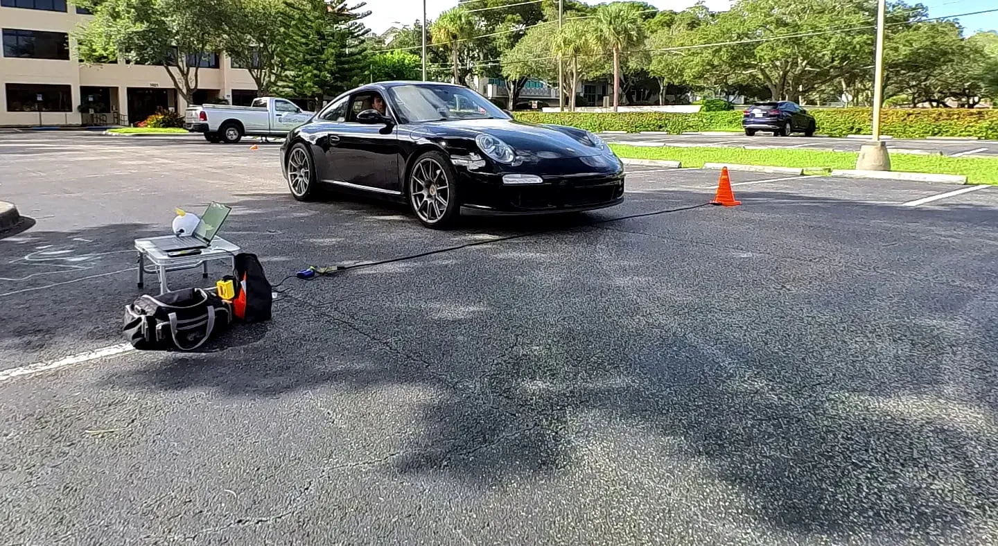 Car 107, a black Porsche 911, rolling up to a pressure tube laid across a car park, with the Timing107 timing box, a laptop and a kit bag beside the tube
